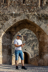 Obraz na płótnie Canvas Man wearing hat and sunglasses standing in the doorway of an old stone castle while checking his cell phone
