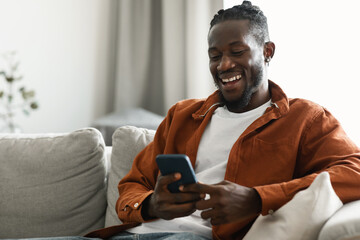 Happy african american man sitting with smartphone in hands and messaging or browsing social...