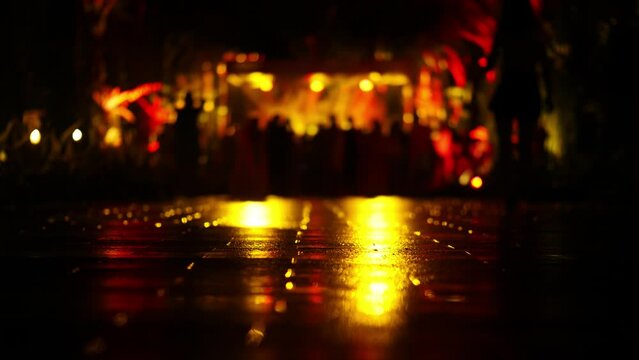 Music performance at night time, out-of-focus view of dancing spectators. Bright flashing lights reflecting at wet pavement, camera focused on the ground