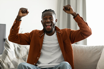 Joyful black man cheering for sports team on television, gesturing YES and shouting, sitting on...