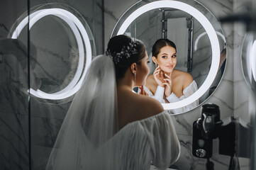 A beautiful, young bride model stands in front of a round mirror with a lamp in the bathroom, reflecting, enjoying the beauty. Wedding portrait, photograph.