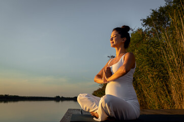 Pregnant woman doing yoga at lake during sunset.