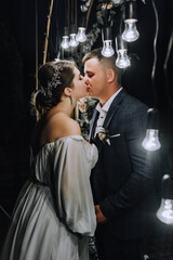 A stylish groom in a blue suit and a beautiful bride in a white dress hug, kiss near an arch decorated with flowers and lamps at a ceremony at night. Wedding photography, portrait of the newlyweds.