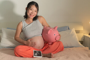 young happy and beautiful Asian Korean woman holding ultrasound and piggy bank sitting on bed...