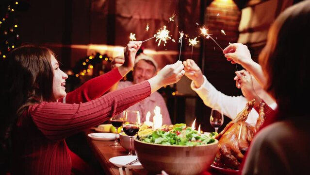 Groups of people celebrate Christmas eve while playing sparkler and have dinner at night which have many food and wine on the table.