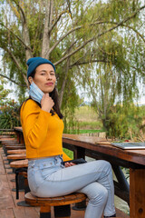 Latin student woman sitting in a park in front of her laptop while removing her protective face mask