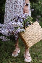 cropped photo of underside young woman in floral dress holds a wicker basket with violet lilac flowers in her hands. Gentle spring atmosphere with fresh, fragrant flowers