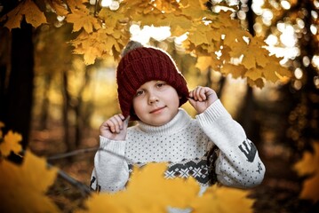 Very smiling boy seven years old in the red wool hat and white sweater is standing in the autumn park behind the yellow maple leaves 