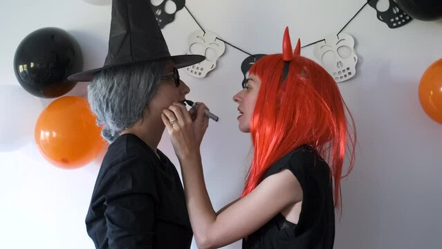 Woman dressed as a demon applying Halloween make-up to her friend dressed as a witch. Halloween costume party.