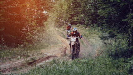 motorcycle racer on enduro sports motorcycle rides on a muddy road with puddles with splashes in forest in an off-road race on sunny day © alexkoral
