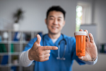 Happy millennial korean male therapist pointing finger at jar of pills in clinic office interior. Treatment with doctor