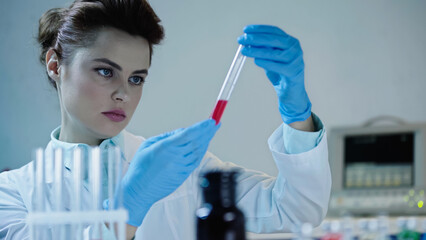 attentive scientist looking at red sample in test tube near blurred medical equipment.