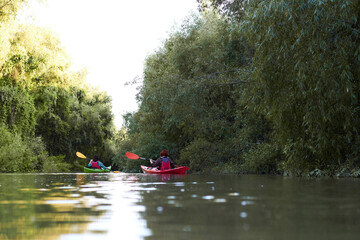 Fototapeta premium Rear view of two women sitting in red and green kayaks. Spring Danube river near trees with gentle green leaves