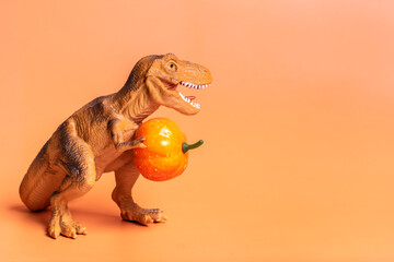 Toy dinosaur Tyrannosaurus holding pumpkin in its paws isolated on orange background Holiday...