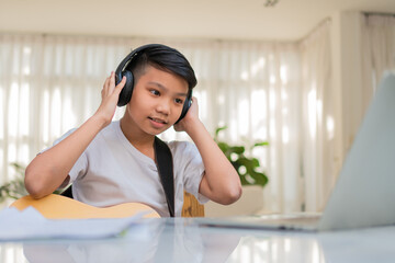 Asian boy playing guitar and watching online course on laptop while practicing for learning music or musical instrument online at home. Boy students study online with video call teachers play guitar.