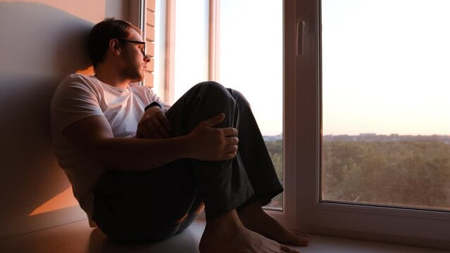 Depressed caucasian man losing job and heartbroken at same time alone, feeling bad and expresses negative. Unhappy attractive young male looks outside window in living room interior, close up.