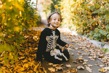 Little toddler boy in halloween skeleton carnival costume sitting on dry leaves in the park in fall. All Saints&#x27; day. School autumn hollidays.