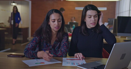 Two diverse female employees working at night in front of computer laptop sitting at office desk talking about work. Young women at their job inside modern workplace in the evening