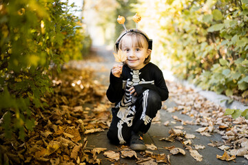Little toddler boy in halloween skeleton carnival costume sitting on dry leaves in the park in...
