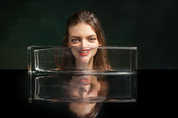 Surreal art photography. Young girl's face through glass vase over dark green background. Object...