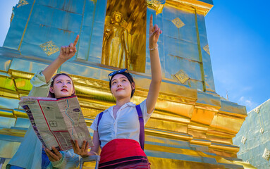 Two women tourists enjoy visiting Thai Buddhist golden temple in Chiang Mai, Thailand. They point out at interesting sites. Vacation and culture concept. 