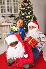Fototapeta na wymiar a family of African Americans - dad, daughter and son in Santa costumes and with gifts in their hands lie on a red plaid and smile against the backdrop of a Christmas tree and decor