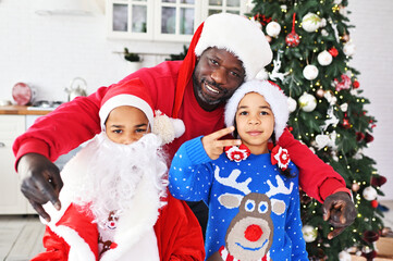 african american family - dad, daughter and son dressed as santa, with a false beard and in christmas sweaters with reindeer smile on the background of the christmas tree