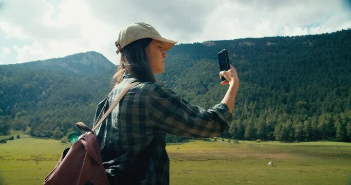Young adult woman on summertime vacation travel make photo using smartphone. Female travel blogger or influencer on mountain trail take picture on mobile phone. People using technology outdoors. 