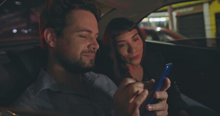 Fototapeta premium Young man showing cellphone screen to female work colleague in car backseat. People riding taxi looking at smartphone device. Person showing content online to friend