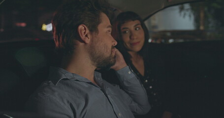 People riding taxi cab in car backseat at night. Young man and woman passengers in the evening after work talking on phone