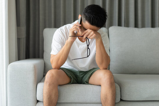 A young Asian man wearing a white T-shirt is talking on the phone and he is feeling stressed on the sofa alone at home.