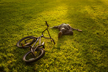 Boy laying on the green grass in sunset light with black stylish bike. Child relax in park