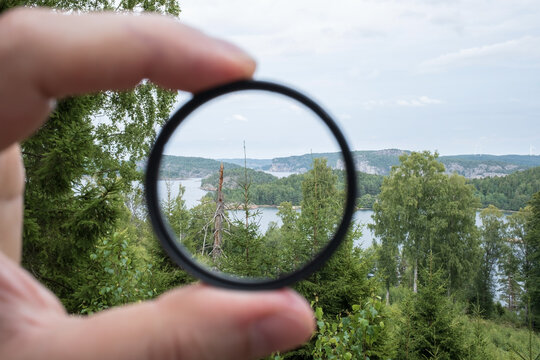 Photographer hand holds a polarizing filter, against the backdrop of trees, sky and sea.