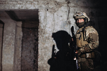 Fototapeta premium Portrait of a Ukrainian defender in the dugout. A soldier with a weapon in his hands casts a shadow on the wall.