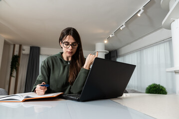 Young college student doing homework at home on her laptop. Girl reading and doing research for her...
