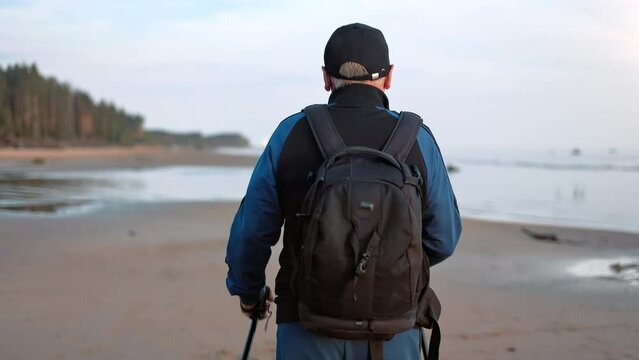 Elderly 70s man backpacker in cap Scandinavian walking stick sport activity at sea beach back view