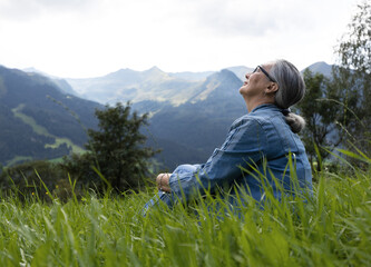 An elderly woman in a denim suit sits on a alpine meadow and looks at the sky