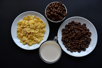 A saucer and a cup of cornflakes and a glass of milk on a black background. Delicious breakfast of cereal with honey and chocolate with milk.