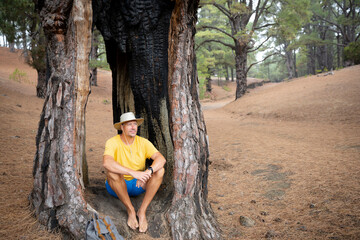 Hiker with hat, yellow shirt and blue shorts sits in the forest on ElHierro in a hollow burnt out tree and takes a break