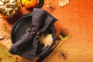 Halloween table setting concept. Black plate, golden cutlery, pumpkins, spooky Halloween decorations and autumn leaves on the table with an orange tablecloth, top view, close up