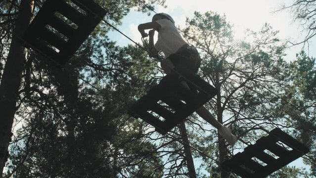 Bottom view brave teenage girl in gear and helmet, overcoming obstacles in a rope park in a sunny wooded area