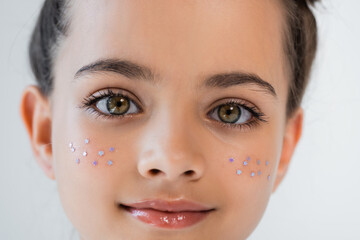 portrait of smiling girl with hazel eyes and sparkling glitter stars on cheeks looking at camera isolated on grey.