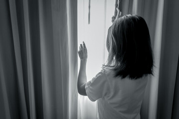 Rear view of woman holding curtain and looking out of the bedroom. Depressed and stressed woman. World mental health day concept. Mental health awarenes. Thoughtful woman in dark room. Unhappy woman.