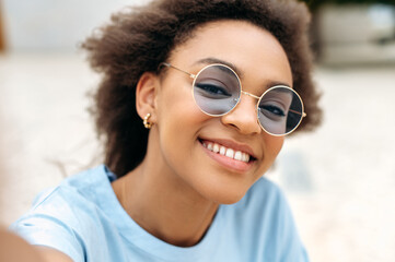 Modern trendy joyful african american girl with curly hair, with blue glasses, video blogger, takes...