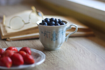 Vintage tea set filled with blueberries, blackberries and starwberries, open book, reading glasses and decorative candles on the table. Selective focus.