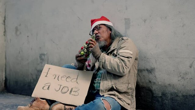 4k, A homeless old Asian man is sitting alone by the wall laughing and drinking, holding a sign asking for help. The madman sat alone on the roadside laughing and smiling.