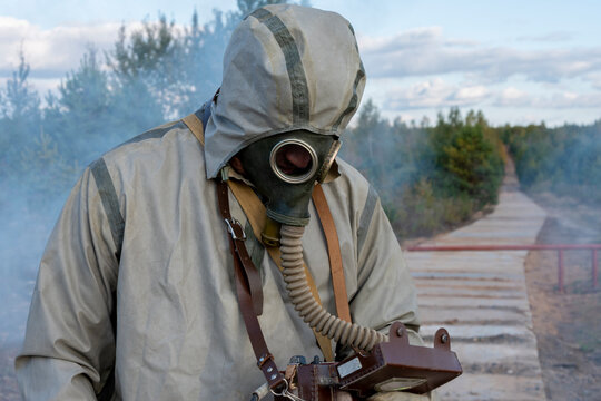 A military man in a chemical protection suit and a gas mask measures the level of radiation on a concrete road, smoke from fires. Concept: consequences of radiation leakage,