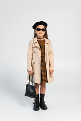 full length of stylish girl in trench coat and beret holding handbag on grey.