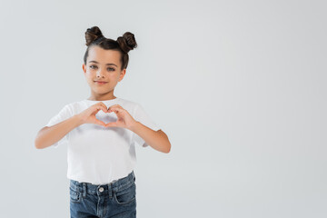 cheerful girl with sparkling glitter stars on cheeks showing heart with hands isolated on grey.