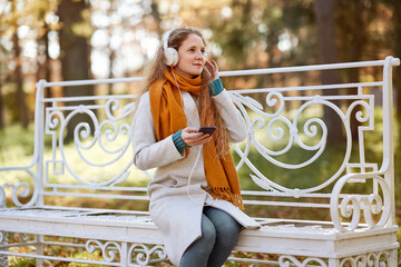 A young red-haired woman is sitting on a bench in an autumn park. A girl listens to music in the...